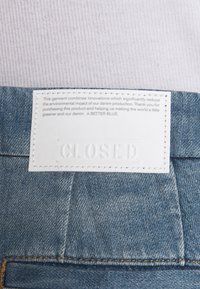 Denimplaggsetikett i vit läder med präglad text "CLOSED." Blå denimbakgrund med synlig sömnad och texturdetaljer.