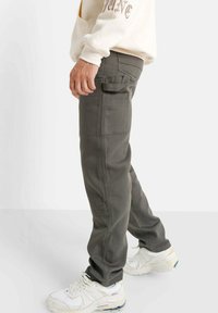 Man in een donkergrijze cargobroek met zijzakken en witte sneakers, staande tegen een eenvoudige lichte achtergrond.