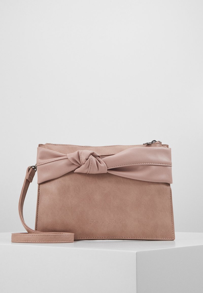 Sac à main rectangulaire rose pâle avec un accent en cuir noué et une bandoulière amovible, posé sur une surface blanche devant un fond uni.