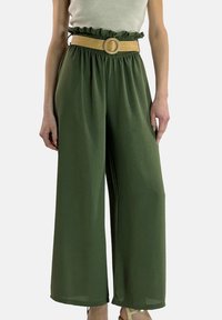 Gröna byxor med vida ben, rynkad midjeresår och ett vävt skärp med rund spänne. Tyget verkar lätt och slätt.