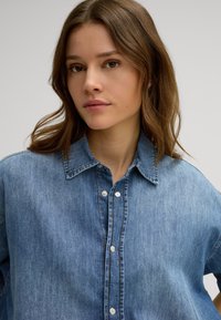 Blue denim button-up shirt featuring a classic collar, metal buttons, and a relaxed fit with subtle texture and stitching details.