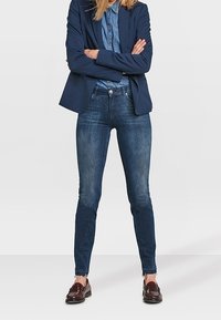 Mujer de pie con los brazos cruzados, vistiendo un blazer azul marino, una camisa de mezclilla, jeans ajustados y mocasines de cuero marrón sobre un fondo simple.