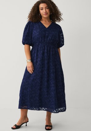 Kvinde med krøllet hår iført en marineblå midikjole af blonder med pufærmer og sorte åbne hæle, poserer foran en ensfarvet baggrund.