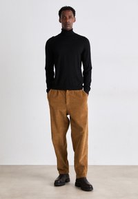 Maglione a collo alto nero abbinato a pantaloni in corduroy marroni. La texture è morbida e i pantaloni presentano una vestibilità rilassata. Stivaletti neri completano il look.