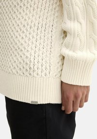 Pletena pulover u boji off-white, sa teksturiranim uzorcima, rebrastim manžetama i donjim rubom, te metalnim logotipom u donjem lijevom kutu.