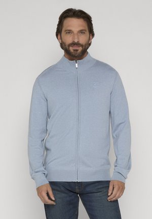 Skæggede mand iført en lyseblå sweatjakke med lynlås, ribkant ved krave og manchetter, kombineret med mørkeblå jeans, stående foran en ensfarvet baggrund.