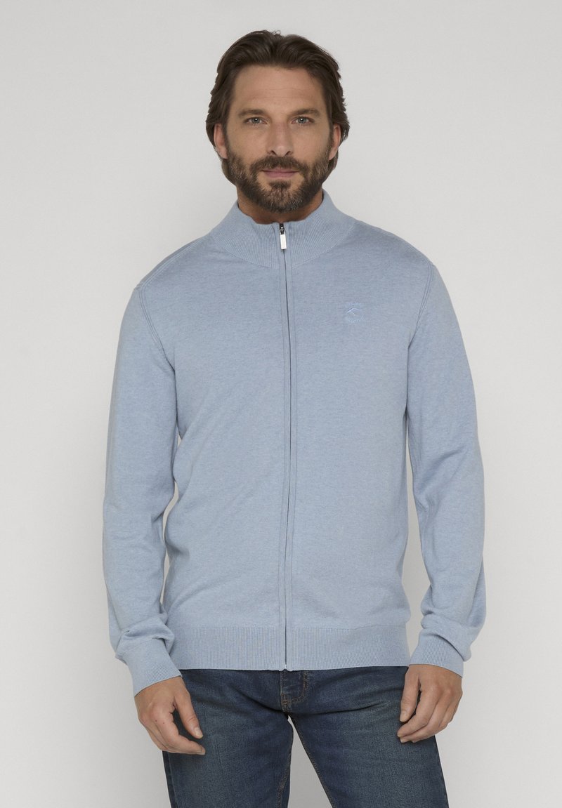 Homme barbu portant un pull zippé bleu clair avec un col et des poignets côtelés, assorti à un jean bleu foncé, debout devant un fond uni.