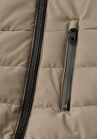 Chaqueta acolchada beige con cremallera negra en el frente y un bolsillo en el pecho con cremallera. Tejido texturizado con secciones acolchadas y detalles de metal negro.