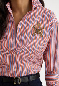 Sieviete, kas valkā sarkanu, baltu un zilu vertikāli svītrainu kreklu ar izšūtu zelta ģerboni uz krūtīm, kombinējot to ar melnu jostu un tumšām biksēm.