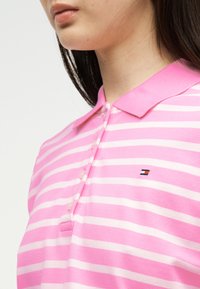 Γυναίκα που φοράει ροζ και λευκή ριγέ πόλο μπλούζα με διπλωμένο γιακά και μικρό κεντημένο λογότυπο στο στήθος.