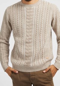 Beige-neulottu neulepusero, jossa on kuvioitu neulos, pyöreä kaula-aukko ja resorit hihansuissa. Alareunan vasemmassa reunassa pieni kolmionmuotoinen logo.