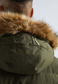 Oliwkowa kurtka z futrzanym kapturem wykończonym jasnobrązowym futrem. Tył zdobią wytłoczone litery "CKT" oraz teksturowany materiał.