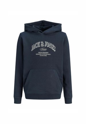 Adolescent aux cheveux bouclés portant un sweat à capuche bleu marine, un pantalon marron et des chaussures beiges, debout et regardant sur le côté.