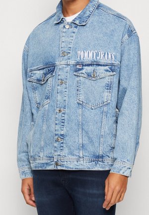 Muž nosí světle modrou džínovou bundu s vyšitým nápisem „TOMMY JEANS“ nad hrudním kapsou, doplněnou tmavými džíny.