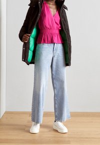Mörkbrun vadderad jacka med grön foder, ljusrosa omslagstopp och vida ljusblå denimjeans. Vita sneakers fulländar outfiten.