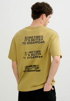 Joven con pelo corto y castaño que lleva una camiseta amarillo mostaza con texto negro "A veces es mejor desaparecer" en la espalda.