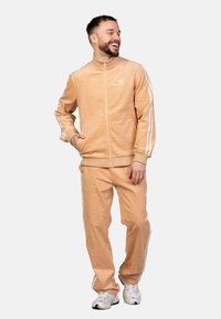 Tuta in corduroy beige con strisce laterali bianche. Presenta un collo alto, chiusura con zip e una vestibilità comoda. Indossato con sneakers bianche.