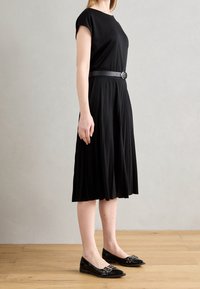 Mustapleatti mekko lyhyillä hihoilla, kapeutettu vyötärö ja sileä pinta. Yhdistetty mustiin ballerinoihin, joissa on koristeellinen hardware.