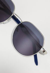 Γυαλιά ηλίου με σκοτεινούς βαθμιαίους φακούς, ασημένιο μεταλλικό σκελετό και μπλε πλαστικά χερούλια; διαθέτουν ένα χαρακτηριστικό ορθογώνιο σχήμα και ελάχιστες λεπτομέρειες.