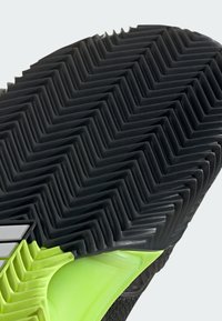 Suela de zapato de goma negra con un patrón en zigzag, que presenta un acento en verde neón en el borde y detalles texturizados visibles para proporcionar tracción.