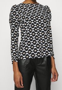 Černý top s dlouhým rukávem s geometrickým vzorem v bílé a béžové barvě. Tkanina vypadá hladce, s nařasenými detaily na ramenou.