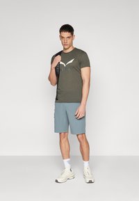 Olijfgroene T-shirt met een wit logo, gecombineerd met lichtblauwe shorts en beige sneakers. De outfit is casual en functioneel.