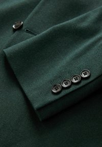 Donkergroen wollen blazer met een gladde textuur, voorzien van een borstzak en vier knopen bij de mouwen met donkere knopen. Aangeschuined ontwerp zichtbaar.