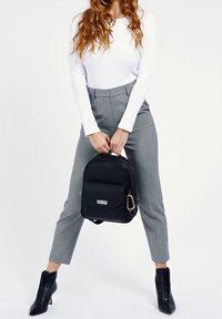 Un sac à dos noir en tissu texturé, avec une poche avant et un accent en métal doré. Associé à un pantalon à carreaux gris et des bottes noires à talons.