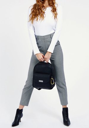 Sort rygsæk med tekstureret stof, der har en frontlomme og et gold hardware-detalje. Sammen med grå ternede bukser og sorte hælshaftstøvler.