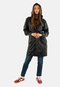Zwarte regenjas met capuchon, voorzien van een gladde textuur en knoopsluiting. Gecombineerd met blauwe jeans en rode sneakers met witte accenten.