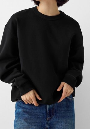 Persoon die een losse zwarte sweater met ronde hals en blauwe spijkerbroek draagt, staand met handen vlak bij de taille tegen een effen achtergrond.