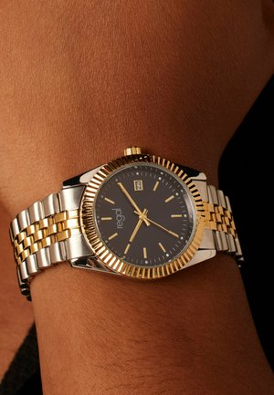 Montre-bracelet bicolore or et argent avec une lunette texturée, un cadran sombre, des index dorés et un affichage de la date à trois heures.