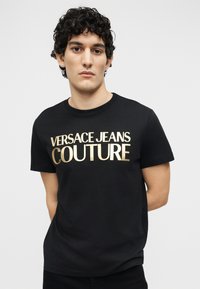 Νέος άνδρας με σγουρά μαλλιά που φοράει μαύρο μπλουζάκι με χρυσό λογότυπο "Versace Jeans Couture", στέκεται μπροστά σε απλό ανοιχτό φόντο.