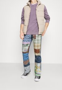 Chaleco acolchado beige sobre un sudadera morada, combinado con pantalones de mezclilla de patchwork que presentan varios patrones, colores y texturas. Zapatillas negras.