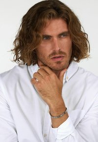 Camicia bianca con bottoni, bracciale d'argento, capelli ondulati di lunghezza media. Mascolinità con una mascella forte e tratti del viso ben definiti, con una texture della pelle morbida e un'espressione neutra.