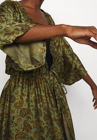 Zielona sukienka w wzory z czarnymi motywami paisley, z szerokimi rękawami, cinchowaną talią oraz zapięciem na wiązanie. Materiał ma gładką strukturę.