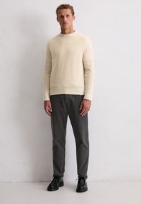 Cremfarbener gerippter Strickpullover kombiniert mit dunkelgrauen cordartigen Hosen und schwarzen Schnürschuhen. Schlichte, strukturierte und figurbetonte Gestaltungselemente.