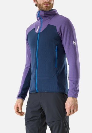 Man wearing a blue and purple zip-up hooded jacket with side pockets and black pants, standing with hands relaxed at sides.