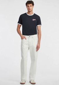 Tumed sinine T-särk valge ääristuse ja roosa "Guess" logoga, kombineeritud heledates, sirgete säärtega teksade ja valgete tennistega, millel on pruunikad aktsendid.