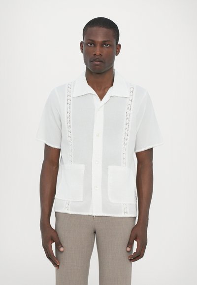Chemise blanche à manches courtes avec un col, ornée de dentelle décorative et de deux poches avant. Tissu lisse, coupe décontractée.
