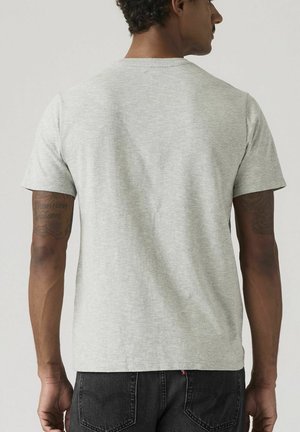 Mand med kort krøllet hår og tatoverede arme iført almindelig lysegrå t-shirts med korte ærmer og mørke jeans, set bagfra mod ensfarvet baggrund.