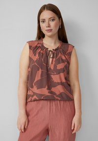 Top sans manches marron et rouille avec un col froncé, présentant un détail à nouer et des motifs abstraits, associé à un pantalon plissé rose pastel.