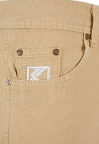 Vaaleanbeige kangaspussi, jossa on kaksi metallista niittiä ja valkoisella brodeerattu logo. Kankaan tekstuuri tuo esiin kankaan rosoisen ilmeen.