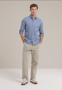 Cămașă albastră deschis cu guler clasic, mâneci lungi; asociată cu pantaloni bej și adidași albi cu accente maronii. Fundal simplu.