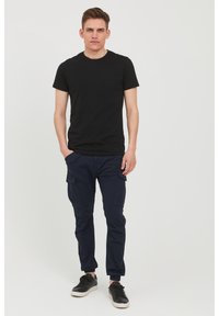 Crna pamučna majica u kombinaciji s tamnoplavim cargo hlačama s bočnim džepovima. Model nosi crne tenisice; jednostavan, ležeran outfit.