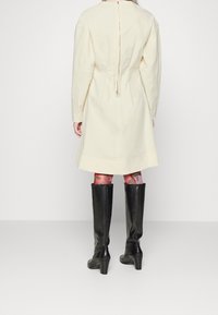 Vestido color crema con mangas largas, terminado con un cierre de cremallera en la parte posterior y un detalle de lazo. La modelo lleva botas negras hasta la rodilla.