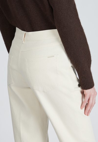 Pantaloni color crema realizzati in tessuto testurizzato, con un taglio dritto. I dettagli notevoli includono due tasche posteriori e una piccola etichetta del marchio.