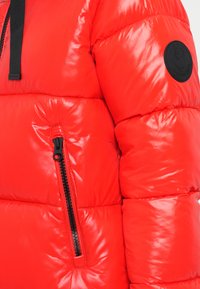 Rote Pufferjacke mit glänzender Oberfläche, ausgestattet mit einem schwarzen Reißverschluss und einem runden, schwarzen Logo-Patch am Oberarm. Hergestellt aus synthetischem Material.