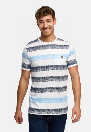 Uśmiechnięty mężczyzna w białej koszulce z teksturowanymi czarno-niebieskimi poziomymi paskami i ciemnoniebieskich dżinsach, stojący na jednolitym tle.