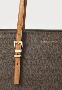 Geantă de umăr maro cu un model, având o suprafață texturată, prezentând o curea din piele bej și accesorii aurii. Logo-ul "Michael Kors" este expus proeminent.